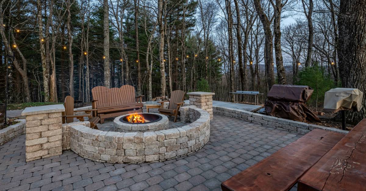  A cozy backyard adorned with wooden seating, stone pavers, and a lit fire pit in the center of the outdoor space.